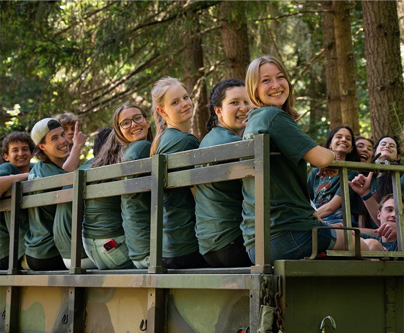 Smiling teens in matching green shirts riding in the back of a truck at camp, surrounded by tall evergreen trees.