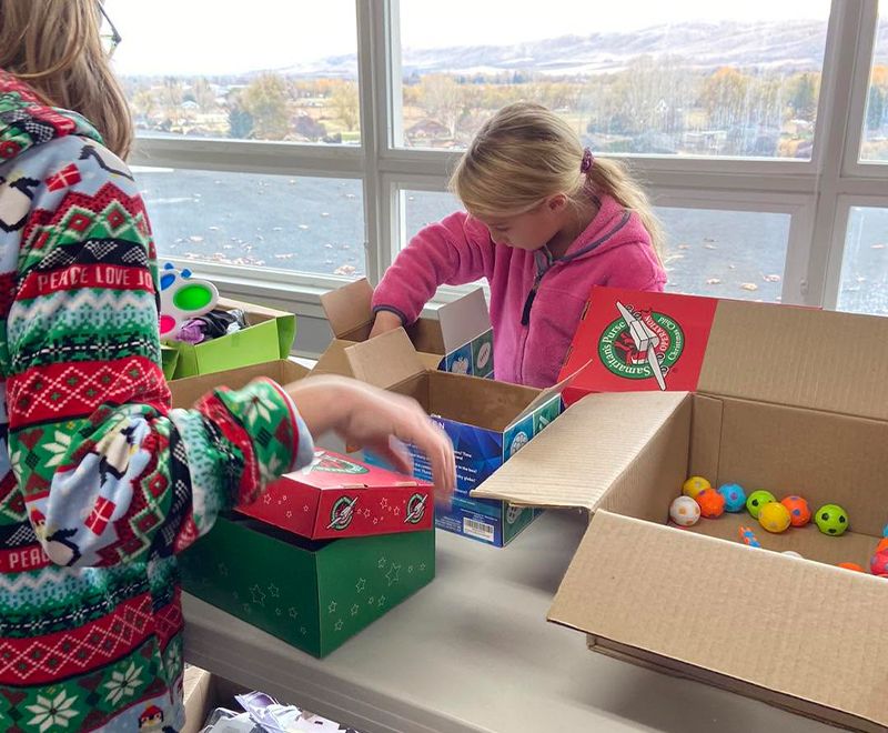 Two children at a table packing shoe boxes with toys, including a box full of colorful balls.